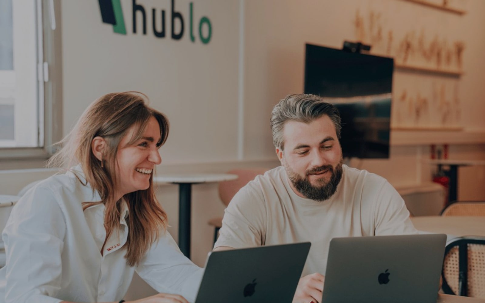 Deux membres de l'équipe Hublo souriant à leurs ordinateurs portables devant le logo de l'entreprise